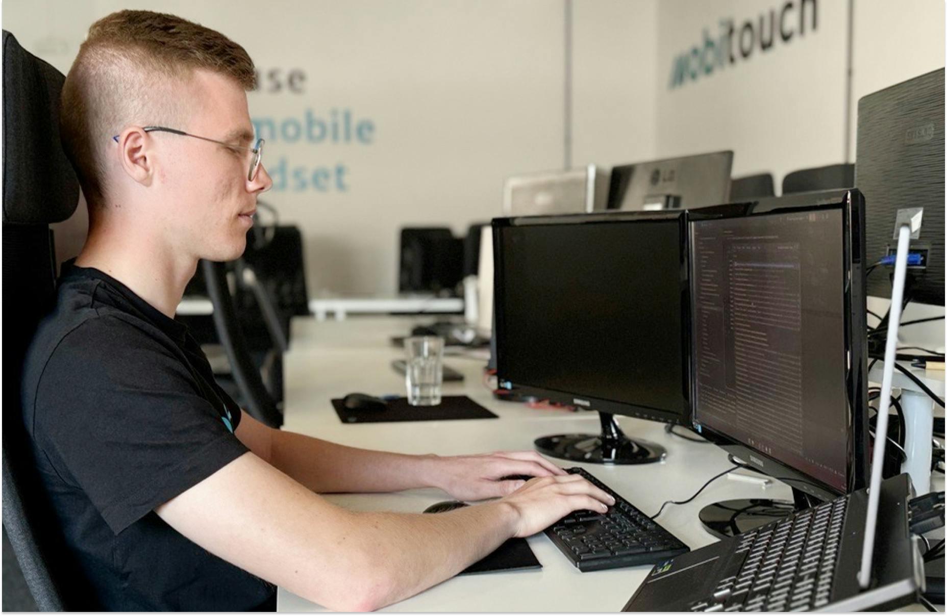 A developer from an IT outsourcing team sits at a desk with multiple monitors, focused on coding